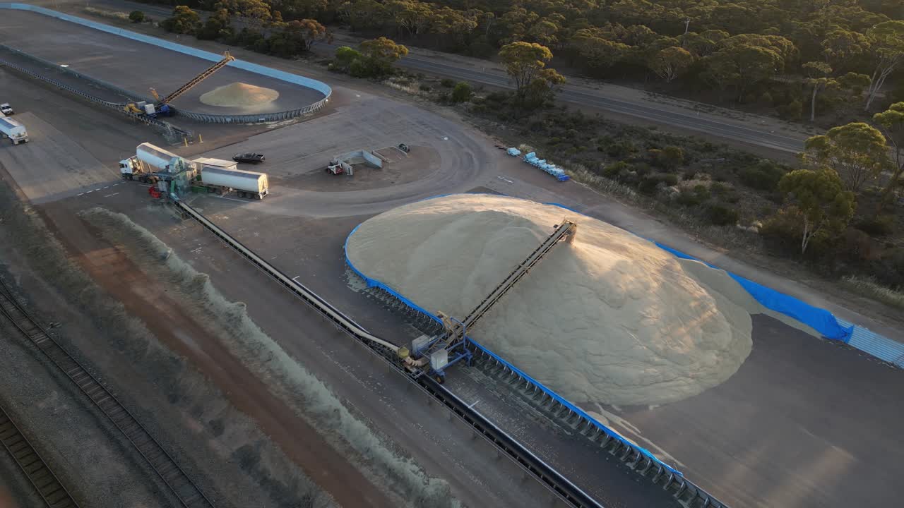 큰 산업 농장 필드의 공중 풍경은 곡물 화물 트럭이 격자에 내리고 해가 지는 동안  컨베이어에서 무더기에 떨어집니다.