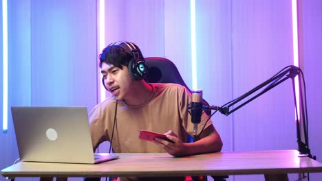 asian man holding mobile phone and read comment in laptop computer then talking to camera while live stream