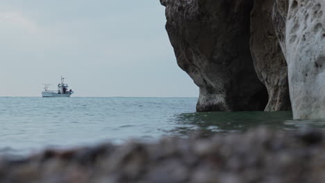 La-Ola-Del-Mar-Se-Rompe-Suavemente-En-La-Cueva-Del-Acantilado-Rocoso,-El-Mar-Mediterráneo-Tranquilo-Con-El-Cielo-Nublado-Y-El-Barco