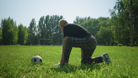 an elderly man kneels on a grassy field, placing his left hand on his leg and his right hand down, with a ball in front of him