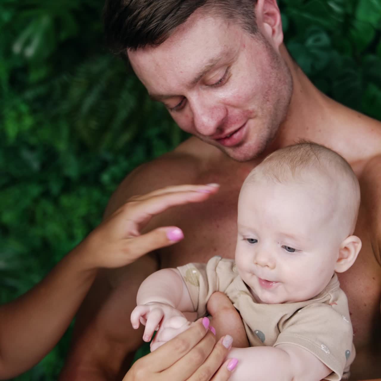 Caucasian dad holding a baby boy in front of him. Female hands clap tiny kid's hands. Vertical view