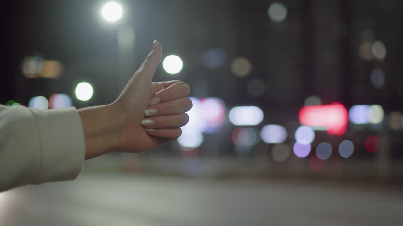 Close-up of woman hand with polished nails wearing white long sleeve giving thumbs up gesture to stop taxi as car passes by on city street at night with colorful bokeh and bright streetlights