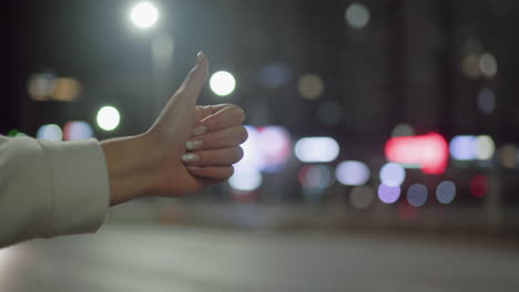 close-up of woman hand with polished nails wearing white long sleeve giving thumbs up gesture to stop taxi as car passes by on city street at night with colorful bokeh and bright streetlights