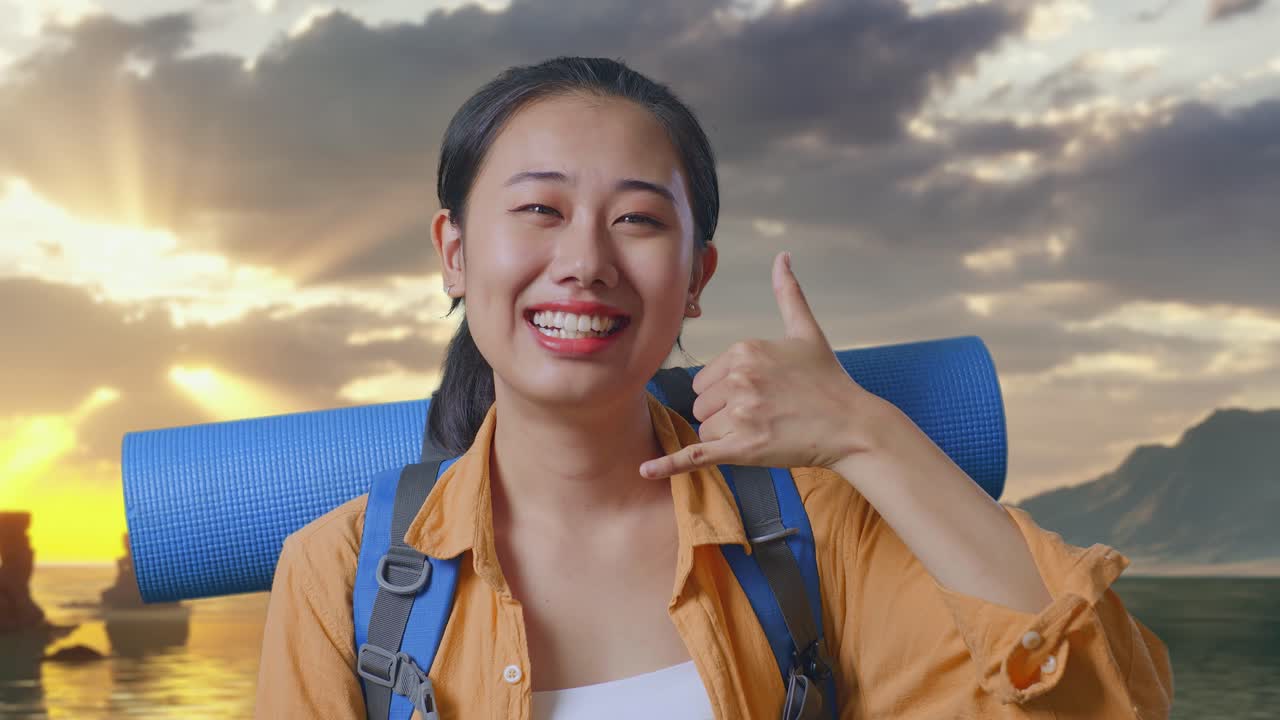 Primo piano di escursionista asiatica con zaino da alpinismo che sorride e fa il gesto "chiamami" mentre si trova sulla spiaggia durante il tramonto