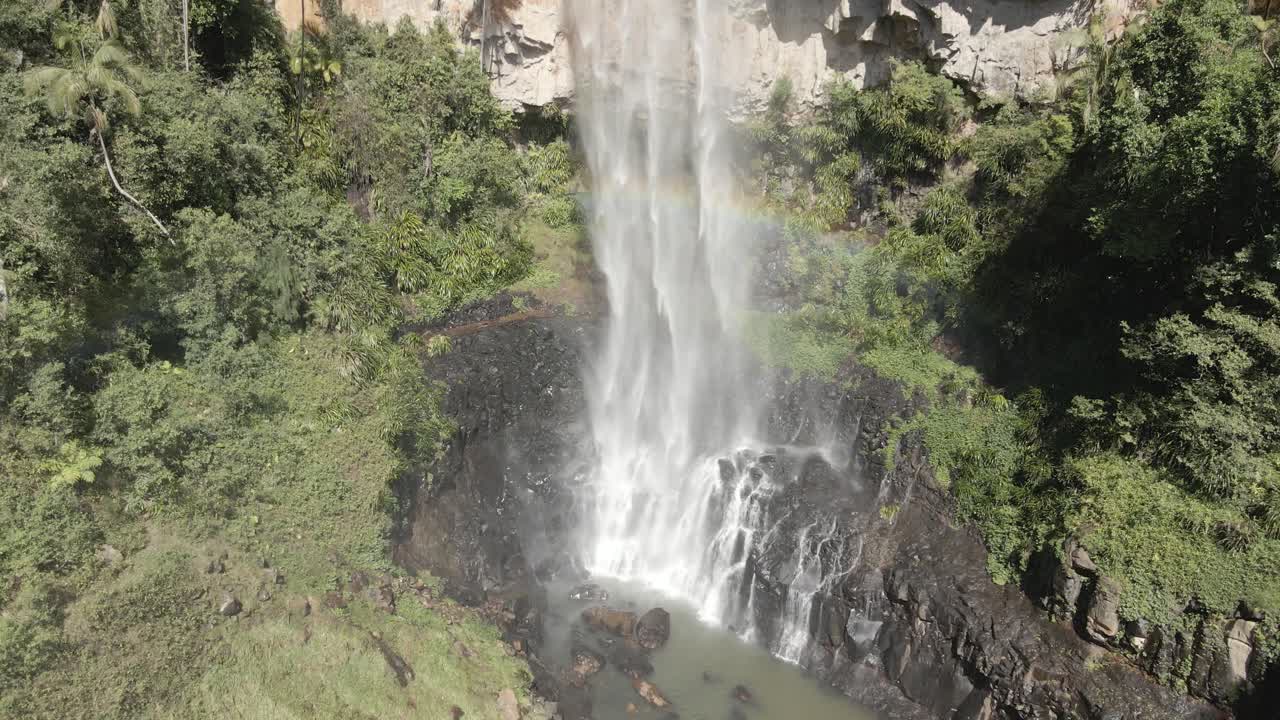 This huge jungle waterfall crashes through lush greenery, mist creating a bright rainbow over the dense landscape. An awe-inspiring display of raw power and tropical beauty.