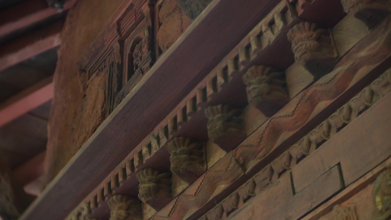 Des boiseries ornent l'intérieur d'un temple tibétain avec des poutres sculptées, des colonnes et des détails de balcon ornementés. Un angle rapproché révèle un artisanat ancien, une lumière chaude et une architecture traditionnelle.