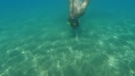 Split-underwater-view-of-fisherman-diving-with-mask-and-yellow-snorkel-looking-for-razor-clams-hidden-in-seafloor-while-holding-bag-full-of-sea-food-with-trabocchi-fishing-platforms-in-background