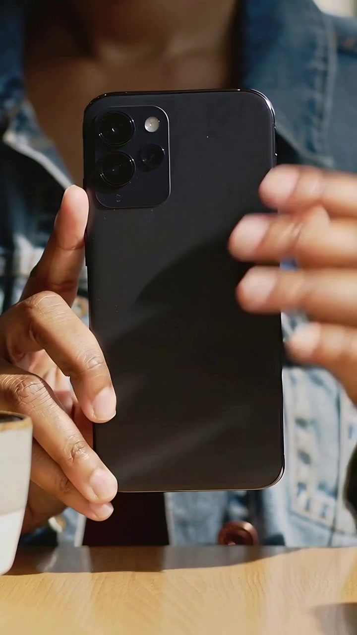 Vertical video: Reaching woman pulling phone from denim jacket at table, showing triple-lens camera