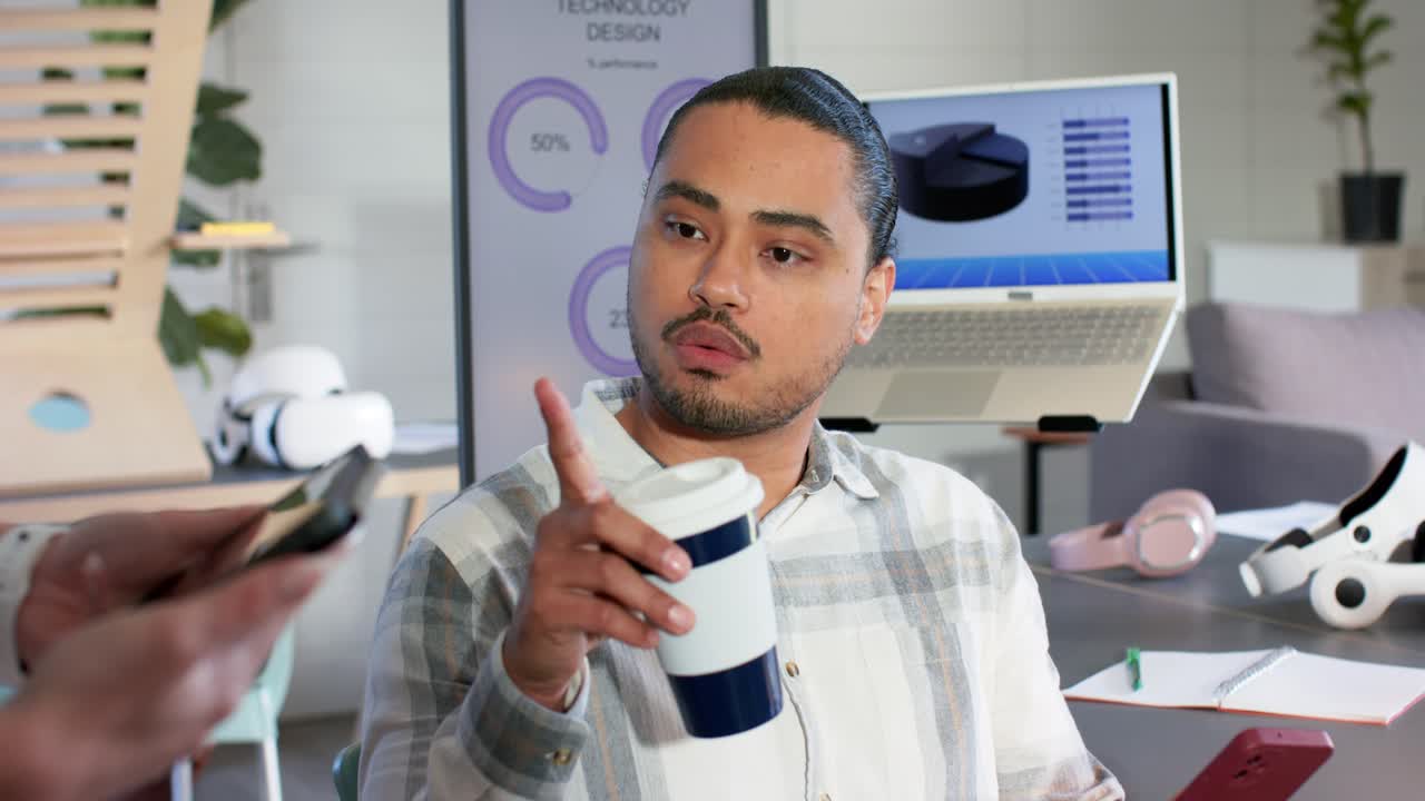 Mid adult man pointing, raising cup while following colleague sharing smartphone and charts at desk