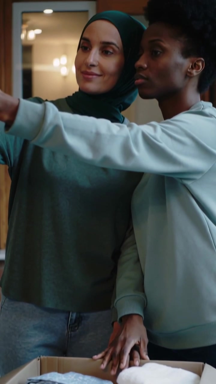 Vertical video: Pulling back at home, two women leaning at box, extending arm for selfie, hijab