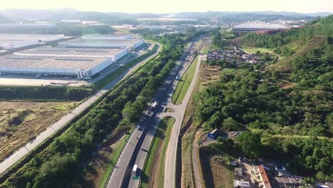 Carretera-Desde-La-Vista-Aérea-Con-Un-Gran-Edificio-Industrial,-Un-Pequeño-Pueblo-Y-Un-Bosque-Montañoso