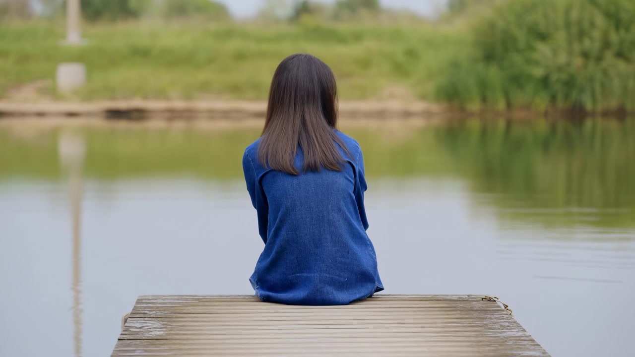 背影顯示一名女子坐在木碼頭盡頭的靜止水面旁，一動不動，柔和的日光、綠色植被和輕柔的倒影構成寧靜的戶外場景
