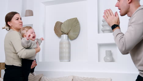 parents are trying to calm down their crying baby and entertain him. mom waves her child while dad makes faces to a kid.