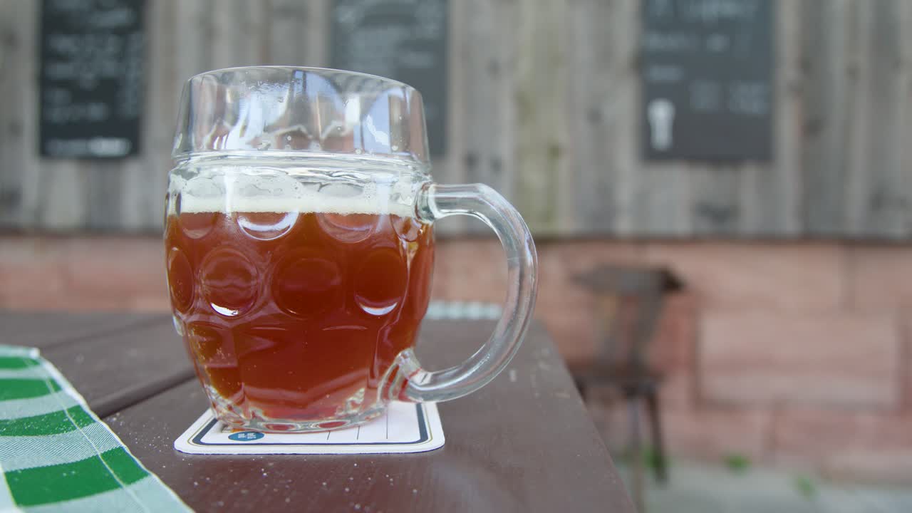 A hand reaches for a traditional beer mug on a wooden table outdoors in daylight