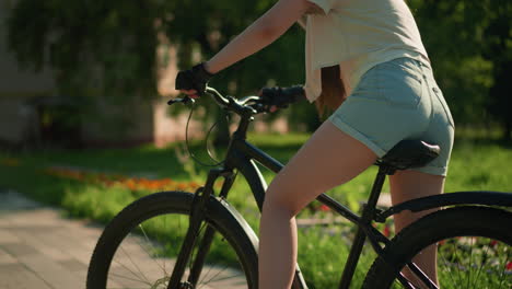 close up of lady mounting bicycle near garden, placing foot on pedal as she prepares for ride, surrounded by colorful flowers, and tall trees with sunlight filtering through, blurred distant building