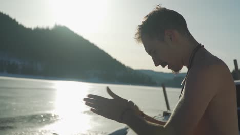 Man,-no-shirt-sitting-in-snow-beside-frozen-lake-wipes-ice-and-cold-water-on-hands,-face-and-chest-to-prepare-for-ice-swimming-polar-bear-plunge