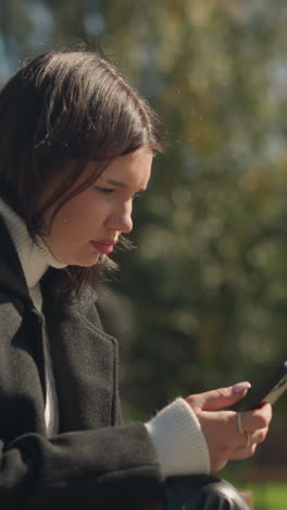 dama sentada al aire libre, centrada en el teléfono en su mano, con el dedo adornado con anillo, y el fondo borroso con vegetación y árboles en un día soleado brillante