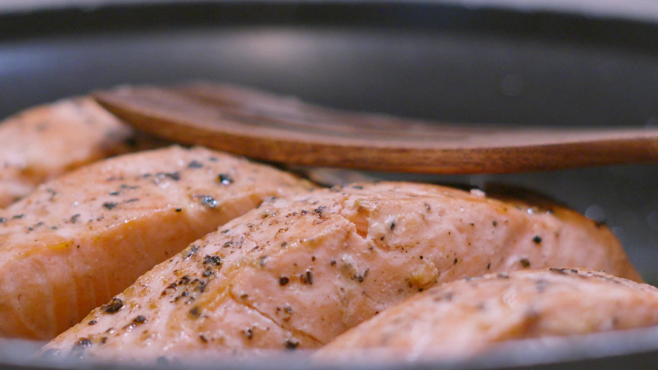 Pomalý pohyb kamery přes ochucené lososové filety na pánvi s dřevěnou lžící položenou nahoře. Vaření s rybami bohatými na Omega-3 pro zdravý životní styl.