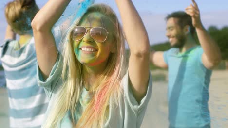 close-up portrait of a beautiful blonde girl dances in celebration of holi festival with her friends. her face and clothes are covered with colorful powder. people dancing in the background.