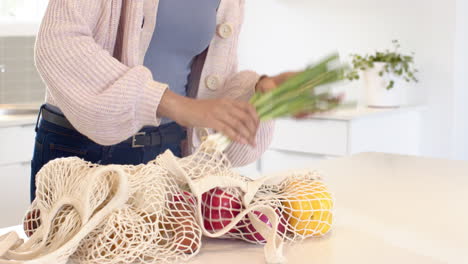 woman in kitchen holding broccoli, unpacking groceries from reusable bag