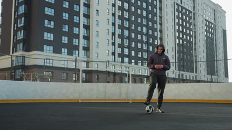 sportsman drops ball to ground, places foot on ball, clasps hands together, and begins turning head, standing confidently in modern urban sports arena surrounded by tall buildings