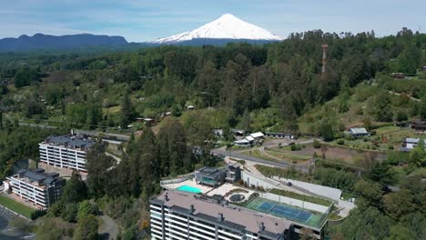 drone backward shot revealing luxurious lakefront condos with pools and tennis court surrounded by lush greenery, framed by the majestic snow-capped villarrica volcano in southern chile