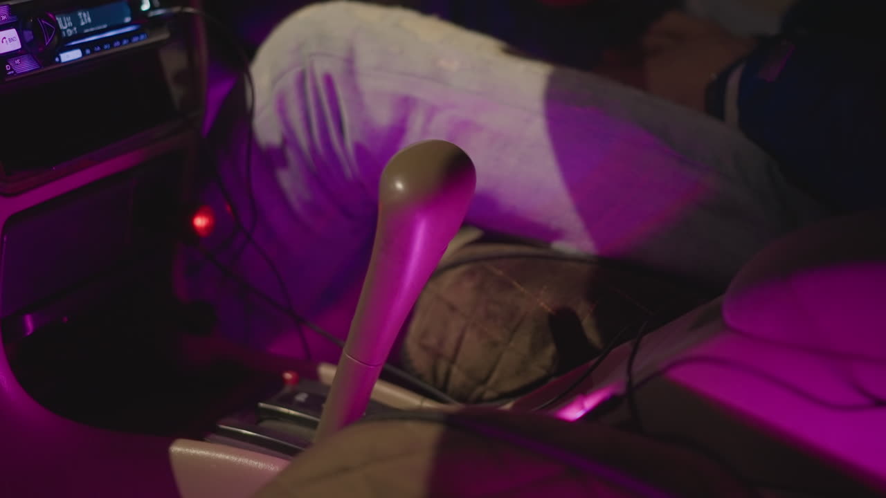 Interior de un coche de noche, mano en la palanca de cambios, vívido resplandor púrpura, movimiento rítmico mientras el conductor se prepara para ir a la discoteca, la muñeca con chaqueta golpea el estéreo, la energía festiva se une al viaje urbano, primer plano de la consola e iluminada