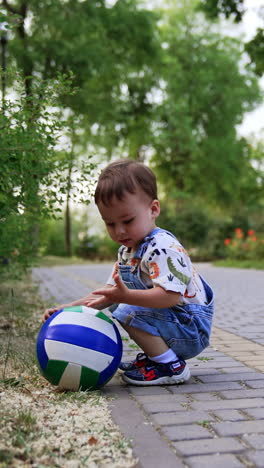 toddler boy in jeans romper wants to lift the ball from the ground. baby misses a little, nearly falls and looks at his palm. vertical video