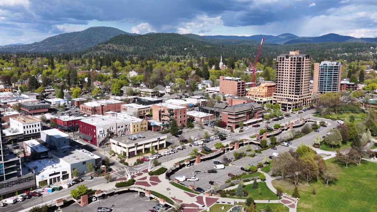 Centro di Coeur d'Alene con moderni grattacieli, affascinanti vetrine in mattoni, parchi verdi, una gru da costruzione e una fitta pineta che si estende verso catene montuose blu in primavera. Ampia ripresa aerea.