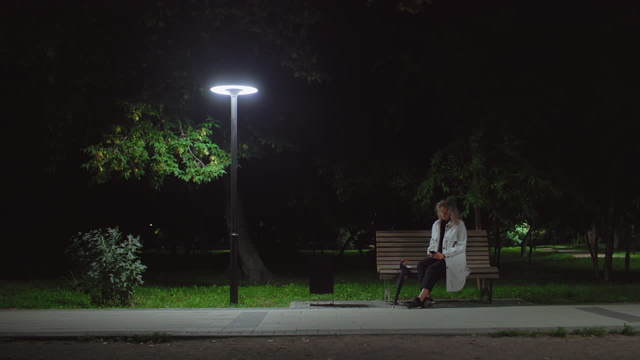 Young girl seated alone on park bench at night operating phone beneath glowing streetlamp, surrounded by quiet park with trees, dressed in white coat, umbrella beside her, distant people walking past