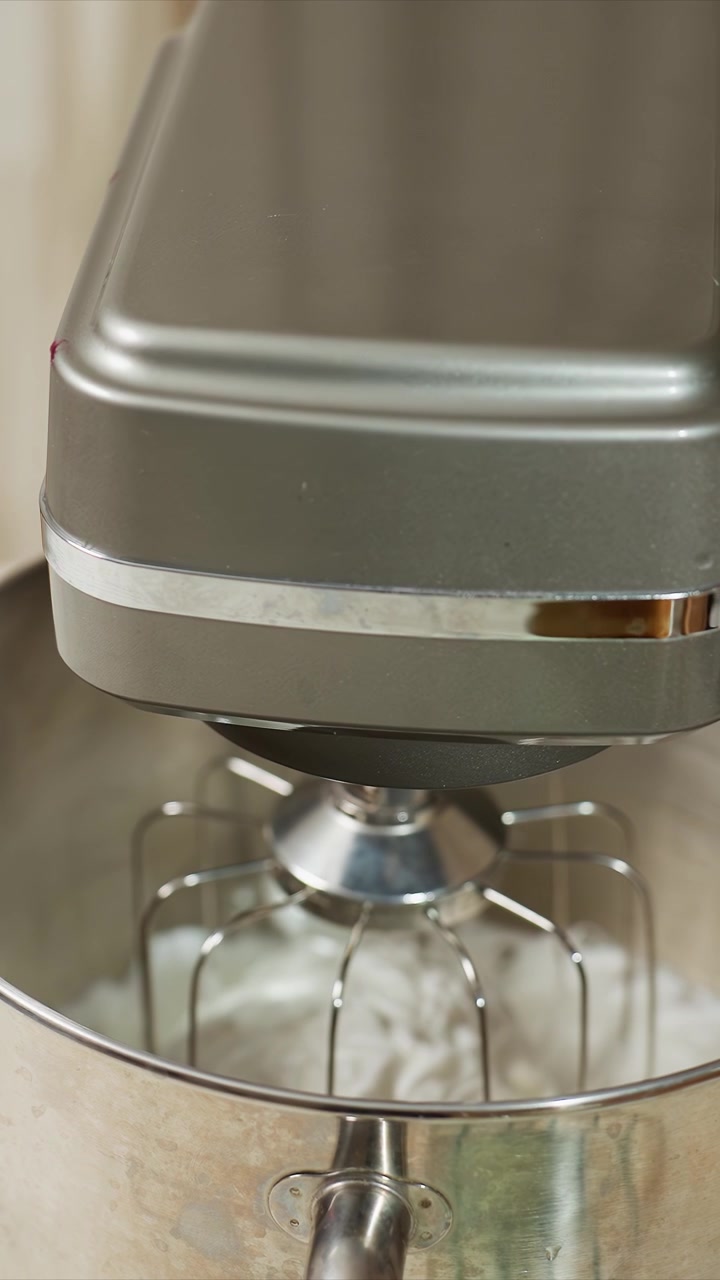 Close up of gray stand mixer mixing white ingredients. Whisk attachment rotates continuously blending contents. Neutral indoor kitchen setting with beige background