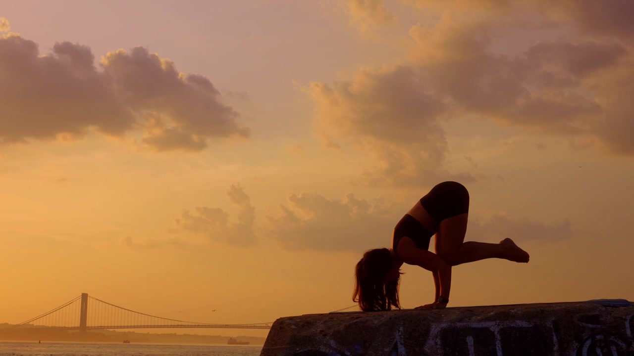 Donna che esegue una posa yoga di squat profondo al tramonto vicino a un ponte costiero. Silhouette di profilo di una praticante su un blocco di pietra vicino all'acqua. Cielo giallo e nuvole sullo sfondo.