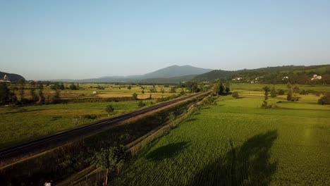 Fliegen-Sie-über-Die-Grüne-Reisfeldwiese-In-Den-Bergwaldhügeln-In-Gilan-In-Der-Nähe-Der-Parallelen-Metalllinie-Der-Eisenbahn,-Des-Tourismus,-Des-Transports-Und-Des-Traditionellen-Landwirtschaftskonzepts-Für-Den-Anbau-In-Der-Erntezeit