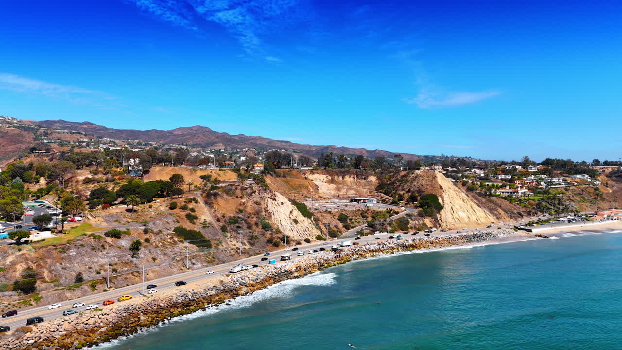 Lively road with many cars parked along the rocky shore of the Pacific Ocean. Aerial perspective on sunny Malibu, Los Angeles County, California, USA