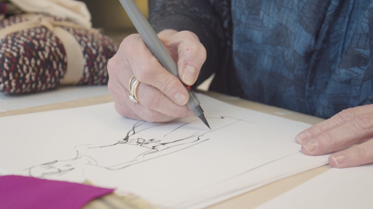 An older woman sketching a woman in a dress, with colored pencils, fabric swatches, and other fashion drawings arranged on her workspace