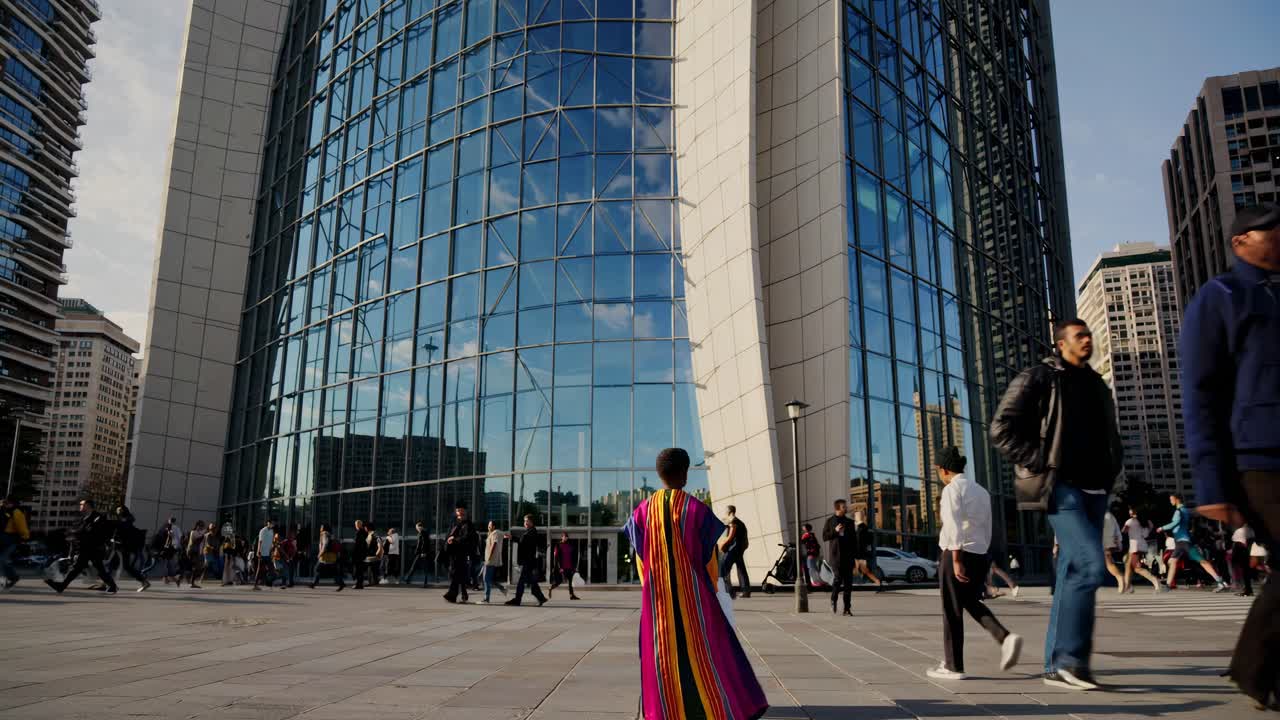 Girl wearing colorful clothes walking towards a modern glass skyscraper in La Defense business district, Paris, France, with people commuting to work