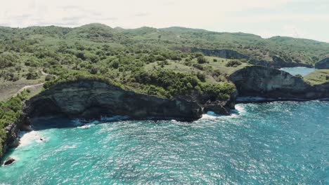 Aerial-drone-shot-of-Nusa-Penida's-beach-in-Bali,-moving-backward-to-reveal-turquoise-waters,-pristine-sands,-and-people-in-the-distance,-framed-by-lush-green-cliffs-for-a-breathtaking-scenic-view