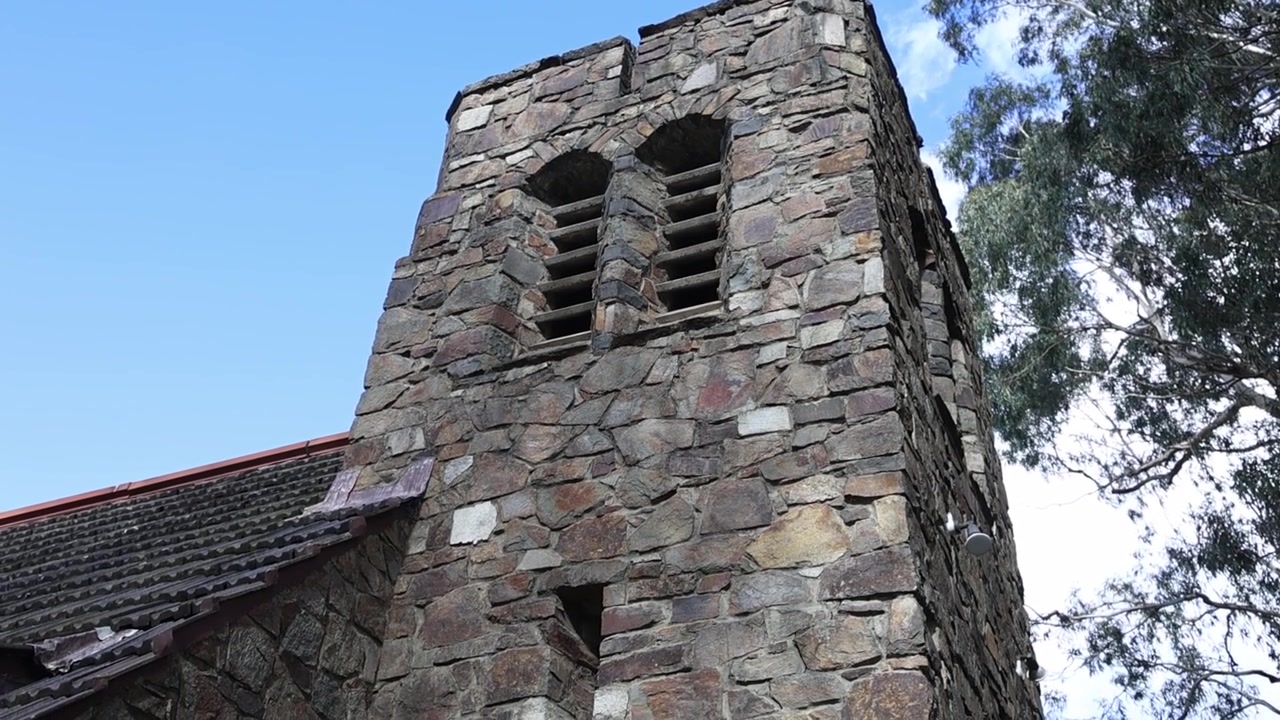Plano aberto para cima de uma antiga torre de igreja de pedra em Olinda, Melbourne, apresentando alvenaria texturizada e design rústico. Rodeada por árvores altas, luz do dia clara e uma atmosfera tranquila e atemporal.