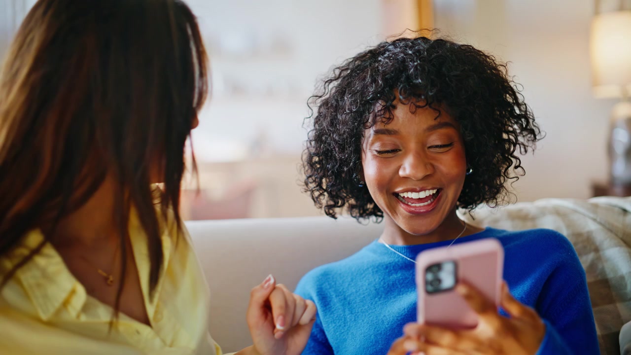 Cheerful girlfriends laughing phone home sofa. Multiracial friends browsing.