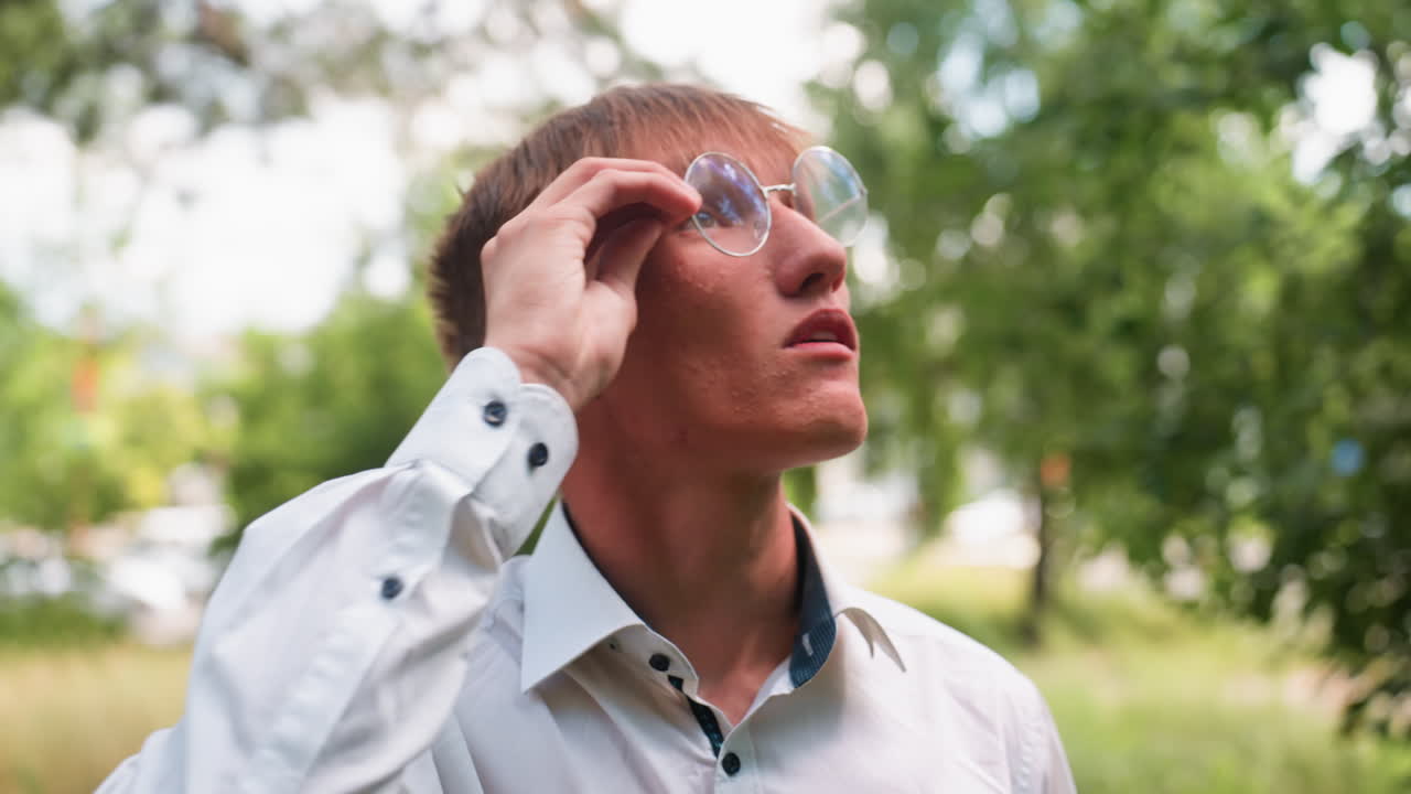 Portrait of young man in white shirt and glasses looking upward while observing trees in outdoor park setting, with blurred background of cars and greenery on bright daylight
