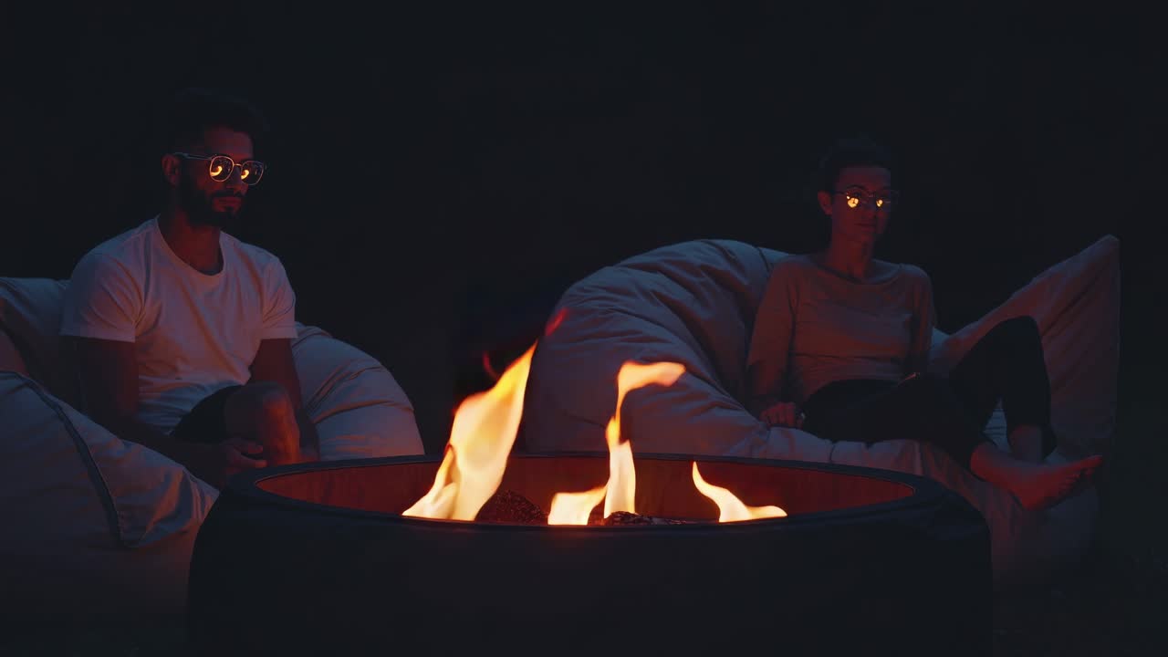 Man and woman relaxing on comfortable beanbag chairs, enjoying a cozy evening by a burning fire pit in the backyard, surrounded by warmth and the soft glow of flickering flames