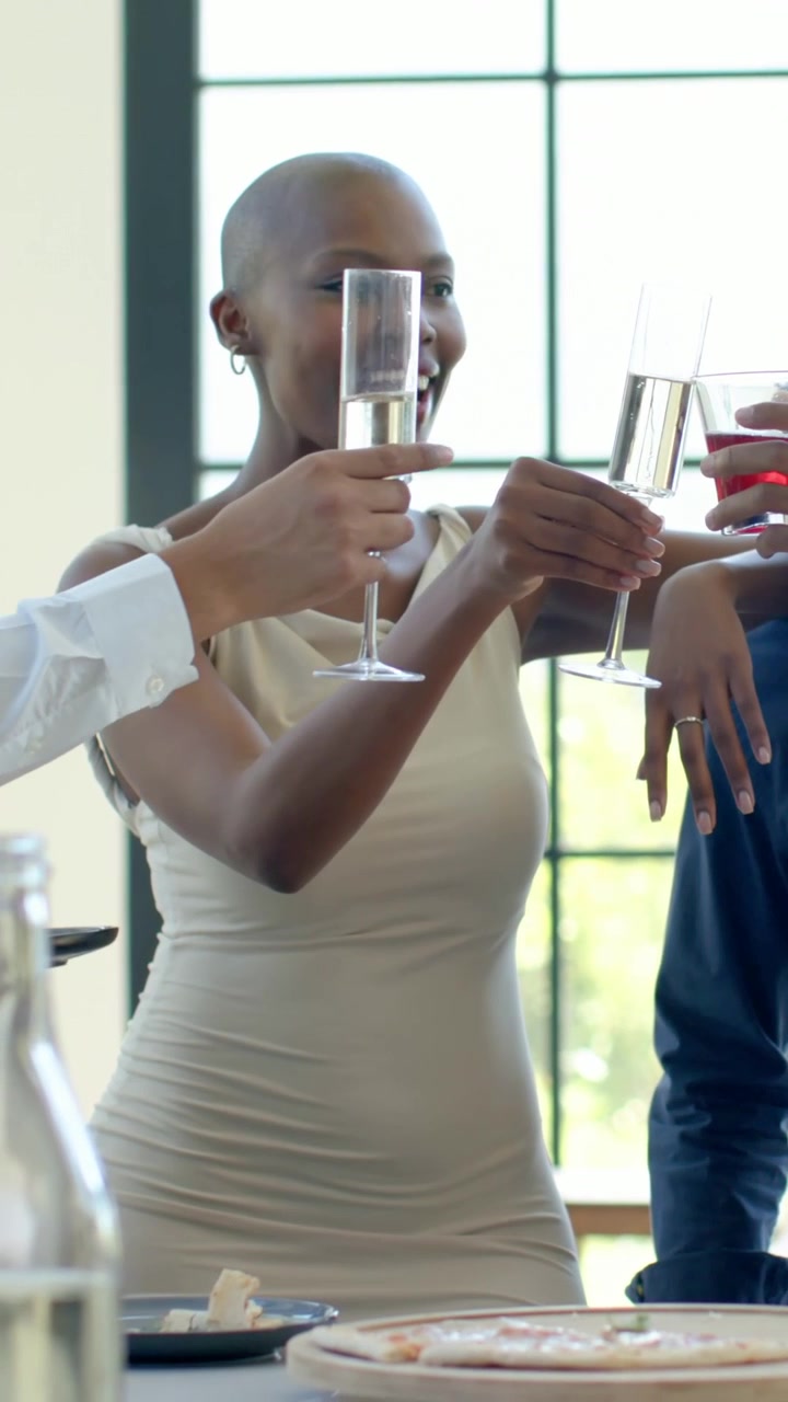 Vertical video: Lifting flute, woman in tan tank dress toasting with flutes at sunlit table, pizza