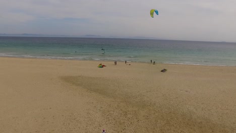 aerial view of a beach with some kite surfers next to the water. one kite is flying high in the sky going side to side and other are on the ground as if they're taking them out. woman walking.