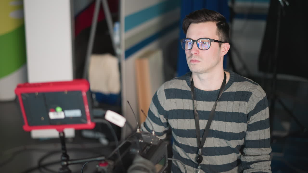 Camera operator intently watching monitor while adjusting camera setup in TV studio. Wearing glasses, focused on equipment as part of filming process, ensuring everything is ready for production