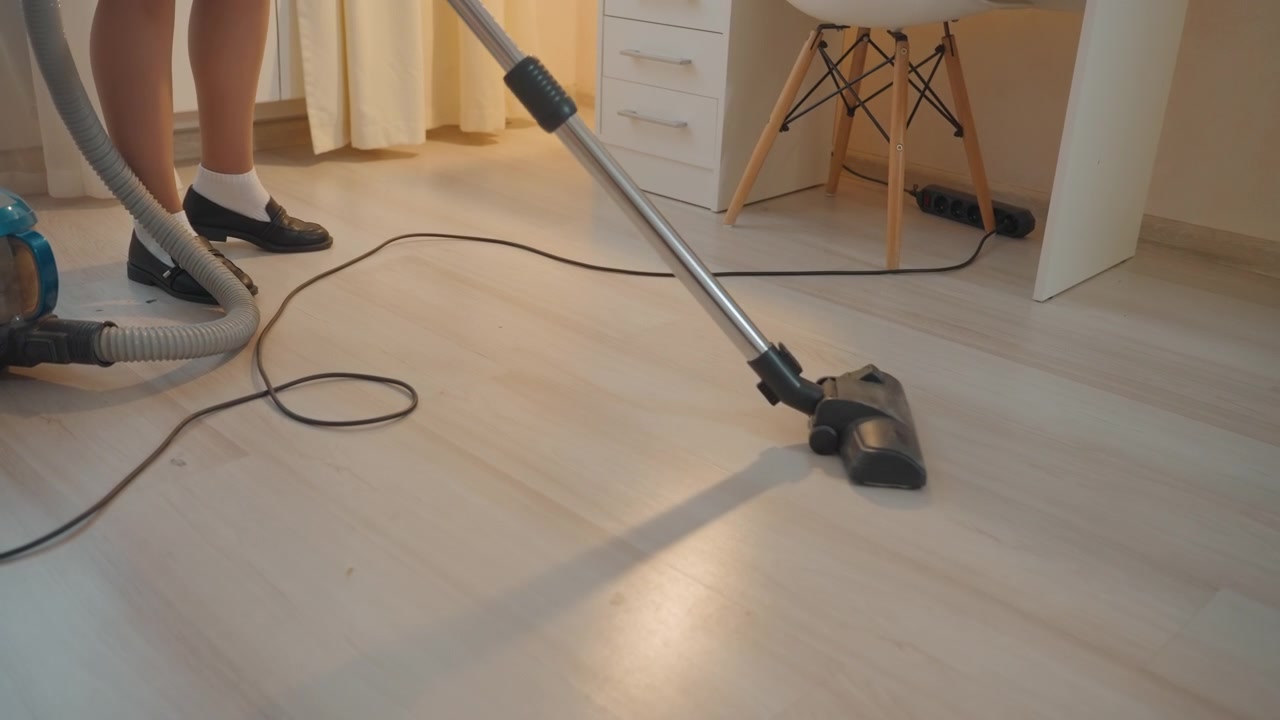 Aspirando piso de madeira dentro de casa iluminada, proprietário move aspirador tipo canister sobre superfície laminada, sala iluminada por cortinas, pernas de cadeira, cabo visível, meias, luz solar suave cria reflexo quente, calmo.