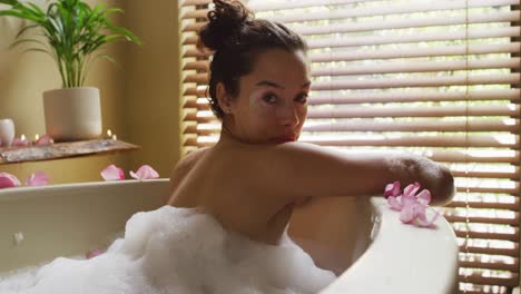 relaxed biracial woman lying in bath with foam and pampering herself with rose petals