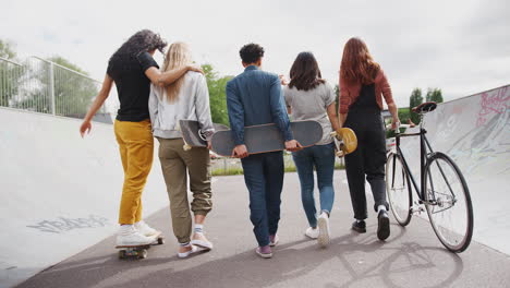 rear view of female friends with skateboards and bike walking through urban skate park