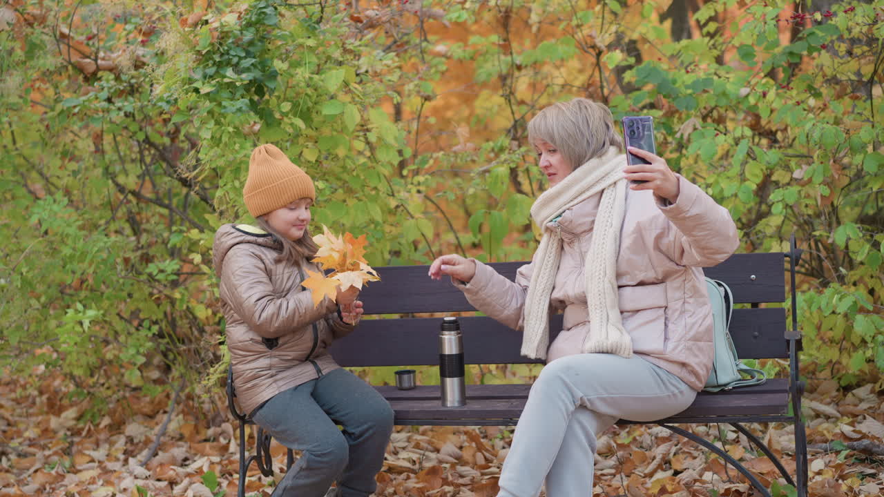 Mother sitting on park bench hands yellow autumn leaves to daughter while taking selfie with smartphone, both dressed in warm jackets and surrounded by vibrant fall foliage and fallen leaves