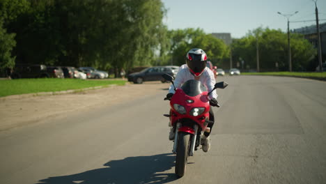 un motociclista con casco monta una bicicleta eléctrica roja rápidamente cerca de un camino de paseo, mientras que un coche rojo se mueve a lo largo, otros vehículos son visibles en la distancia con un poste eléctrico al lado