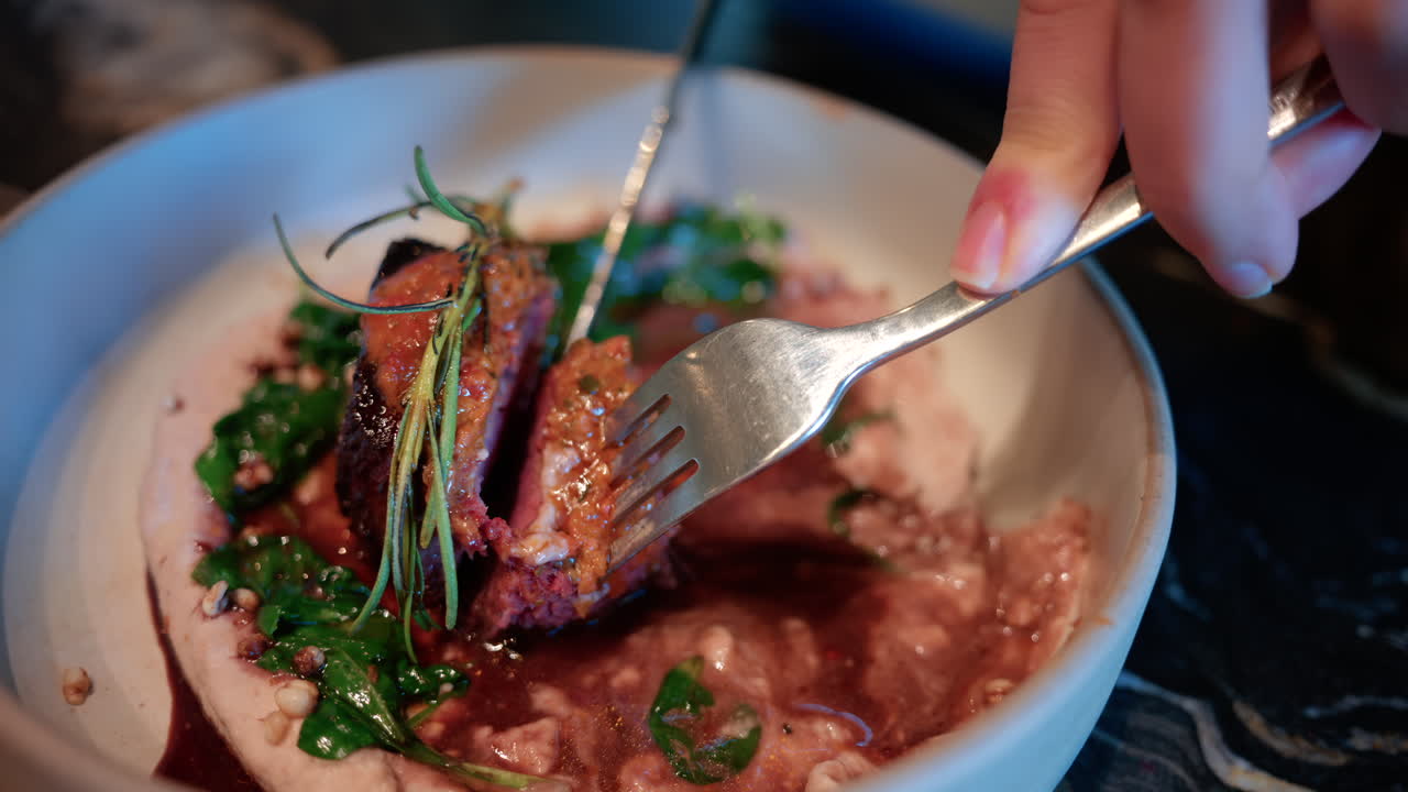 Close up of a woman's hand cutting into a gourmet meat dish served with sauce, greens, and rosemary garnish on a white plate